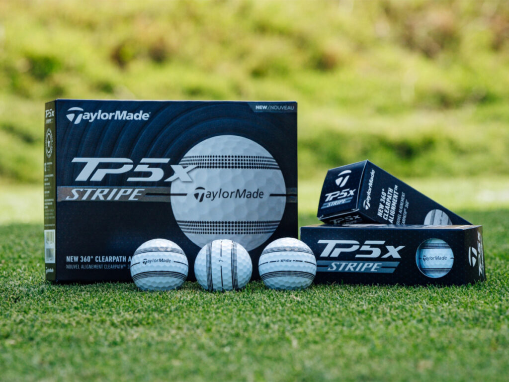 Some TaylorMade TP5x Stripe golf balls are sitting on grass in front of their packaging.